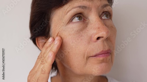 Close-up of mature woman gently touching the skin under her eye, showing wrinkles and puffiness on a light background. Visible signs of aging, natural beauty, slow motion