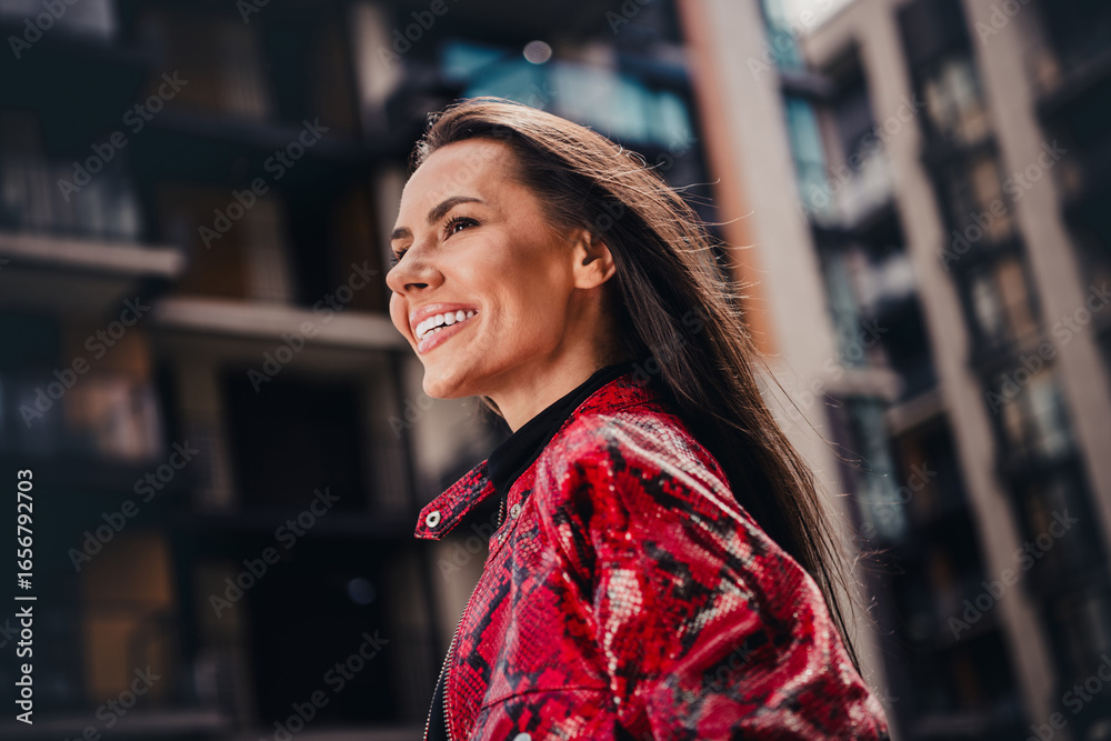 Fototapeta premium Charming young woman in vibrant red jacket smiling outdoors in urban street setting during daylight