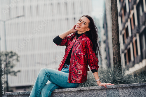 Stylish woman in red jacket enjoying a leisurely moment outdoors in an urban city setting on a sunny day