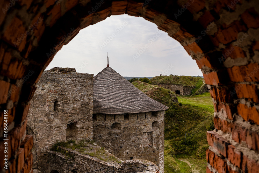 Fototapeta premium Medieval Kamianets-Podilskyi fortress viewed through an archway, showcasing stone walls, grassy landscape, and a thatched roof, embodying historical architecture and cultural heritage