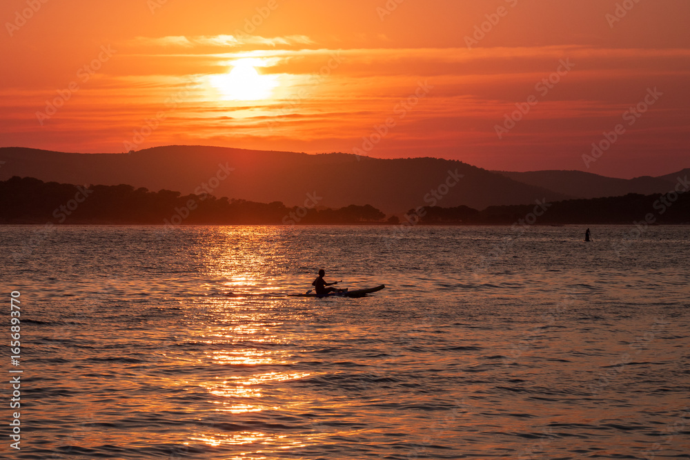 Fototapeta premium Kayaker on the Adriatic Sea during sunset in Biograd na Moru, Croatia. Peaceful seascape with warm orange light and silhouette, ideal for wall art and relaxation themes.