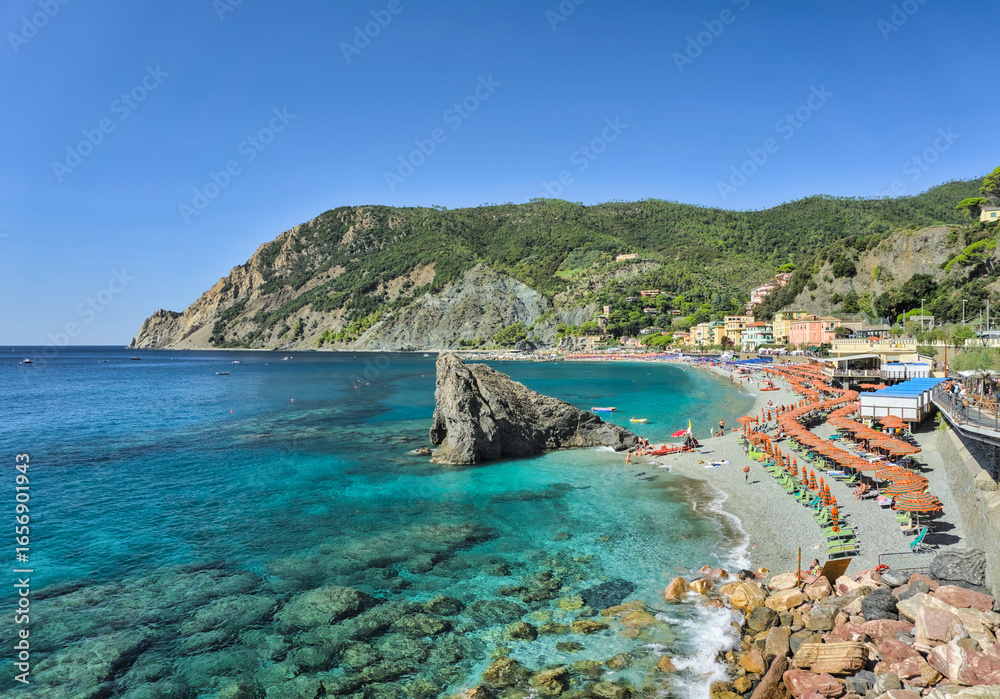 Obraz premium A view of the main beach at Monterosso al Mare, showing the vibrant turquoise water, a prominent rock formation, and a sandy shore filled with sunbathers and beach chairs