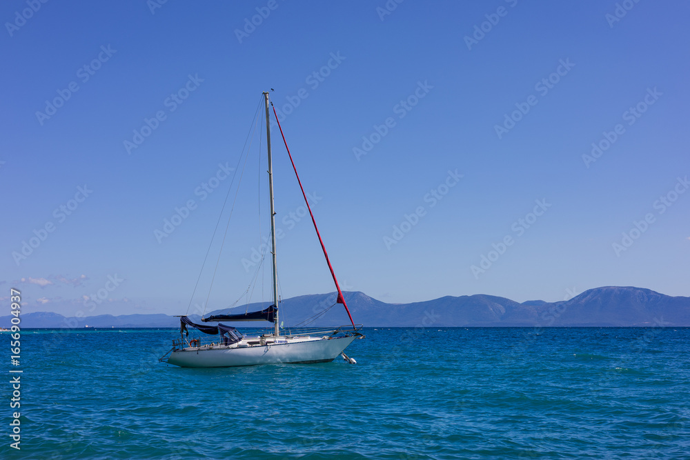 Fototapeta premium A sailing yacht in a calm sea against a backdrop of rocky shores in the rays of bright summer sun. Peaceful seascape. Adriatic Sea, Croatia.