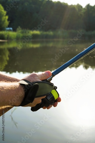 A close-up shot of a person holding a fishing rod, with a visible fishing reel and bright green fishing line. The photo is taken outdoors by a peaceful body of water with trees in the background.