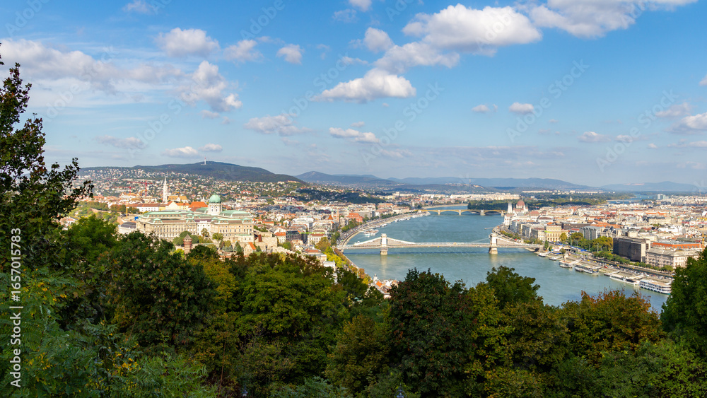 Fototapeta premium Panoramic view from Gellert Hill lookout showing Danube River's curve, Parliament building, Castle Hill with historic rooftops, and green hills surrounding Budapest. Budapest, Hungary.