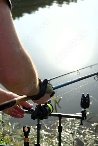 A close-up image of a person adjusting a fishing rod by the water, preparing to fish. The photo shows the fishing reel and line, with fishing rods set up in a peaceful outdoor setting by a lake.