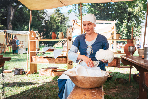 Valokuva Medieval woman in traditional dress preparing bread dough during historical reenactment event outdoors, surrounded by wooden table, clay pottery and rustic baskets under festival tent