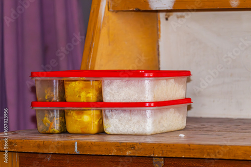 Freshly packed homemade meal for hostel with rice and curry in red-lid plastic boxes on wooden desk.