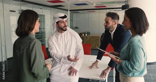 Young multicultural teammates engaged in briefing in meeting room. Arabic man dressed in traditional kandura lead professional discussion with Latin colleagues. Teamwork, ideas exchange, leadership