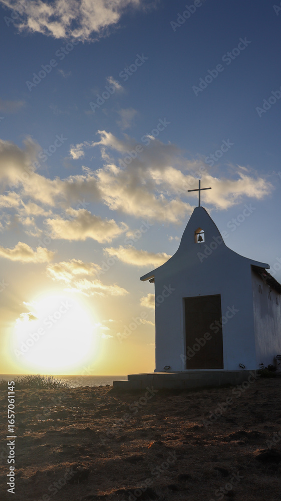 Fototapeta premium Amanhecer na Capela de São Pedro em Noronha, Brasil. O sol nasce refletindo no Atlântico e no pequeno templo histórico que simboliza fé e tradição pesqueira