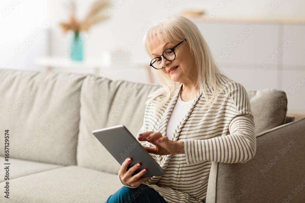 Portrait of mature lady wearing glasses spending time with digital tablet at home, browsing internet, watching videos, checking social networks while relaxing on sofa in living room, free copy space Portrait of mature lady wearing glasses spending time with digital tablet at home, browsing internet, watching videos, checking social networks while relaxing on sofa in living room, free copy space