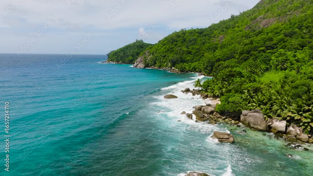 Waves crash against large rocks on the tropical coastline, surrounded by dense green foliage. Seychelles, Mahe. Waves crash against large rocks on the tropical coastline, surrounded by dense green foliage. Seychelles, Mahe.
