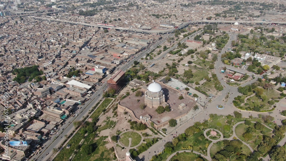 Drone view showing a famous Sufi shrine with a white dome surrounded by gardens and courtyards, set against the dense urban sprawl of Multan city. Drone view showing a famous Sufi shrine with a white dome surrounded by gardens and courtyards, set against the dense urban sprawl of Multan city.