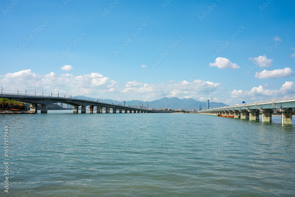 Two long bridges span the calm waters leading to the urban cityscape and distant mountains under the clear blue sky of Xiamen. Two long bridges span the calm waters leading to the urban cityscape and distant mountains under the clear blue sky of Xiamen.
