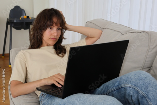 Tired young beautiful curly brunette sitting on the sofa and working at the laptop at home