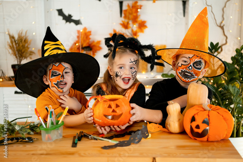 Halloween, kids boys and a girl in festive costumes and makeup are preparing for Halloween at home in the kitchen by coloring and carving pumpkins and scaring everyone