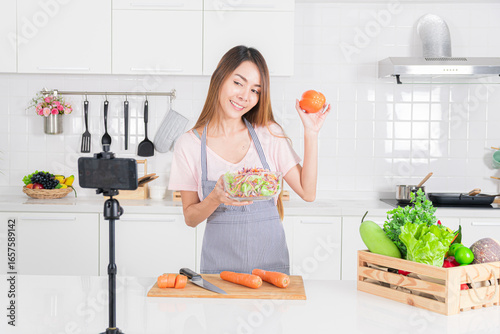 Engaging woman enthusiastically show freshly made salad and capsicum to her smartphone camera, creating content about healthy eating in her bright kitchen.
