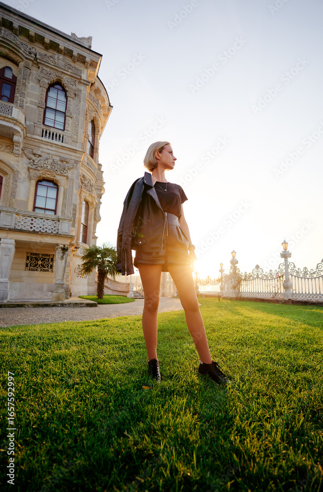 Fototapeta premium A stylish woman poses confidently on a grassy lawn with a historic building in the background during a warm sunset, creating a vibrant yet serene ambiance.