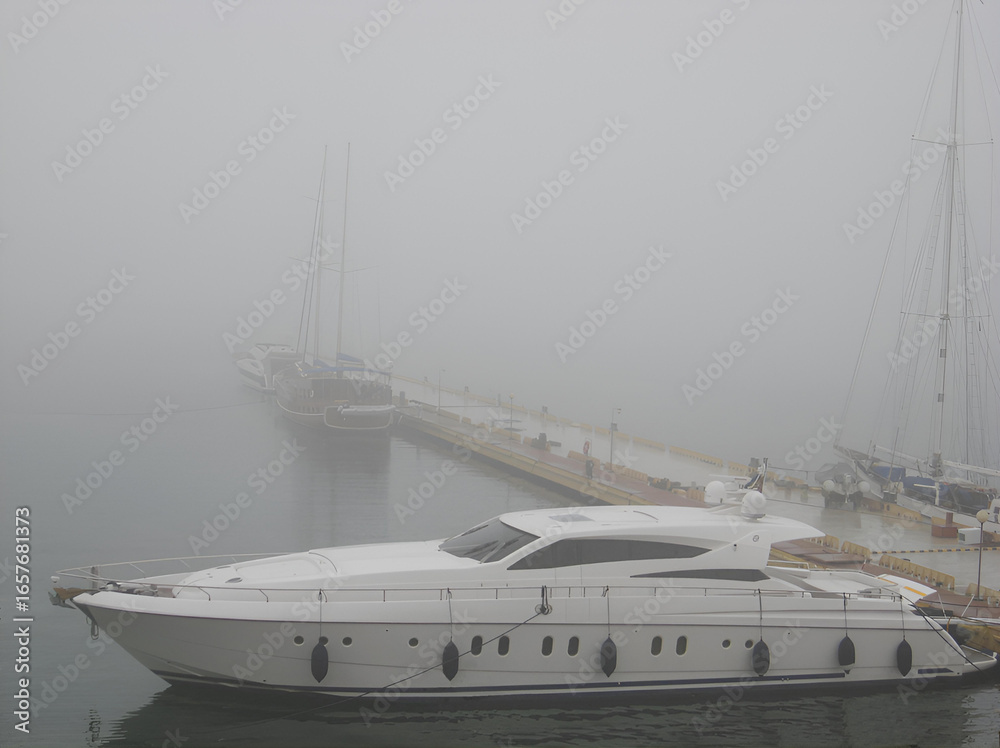 Fototapeta premium Elegant yacht docked at pier on a foggy morning, sailing boats hidden in mist, tranquil seascape symbolizing mystery, luxury travel, marine leisure and nautical atmosphere