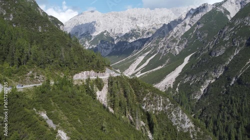 Aerial view of Hahntenjoch valley near Imst, Austria, with winding alpine road, rocky slopes, lush green patches, and dramatic mountains under a partly cloudy sky, showcasing rugged natural beauty.
