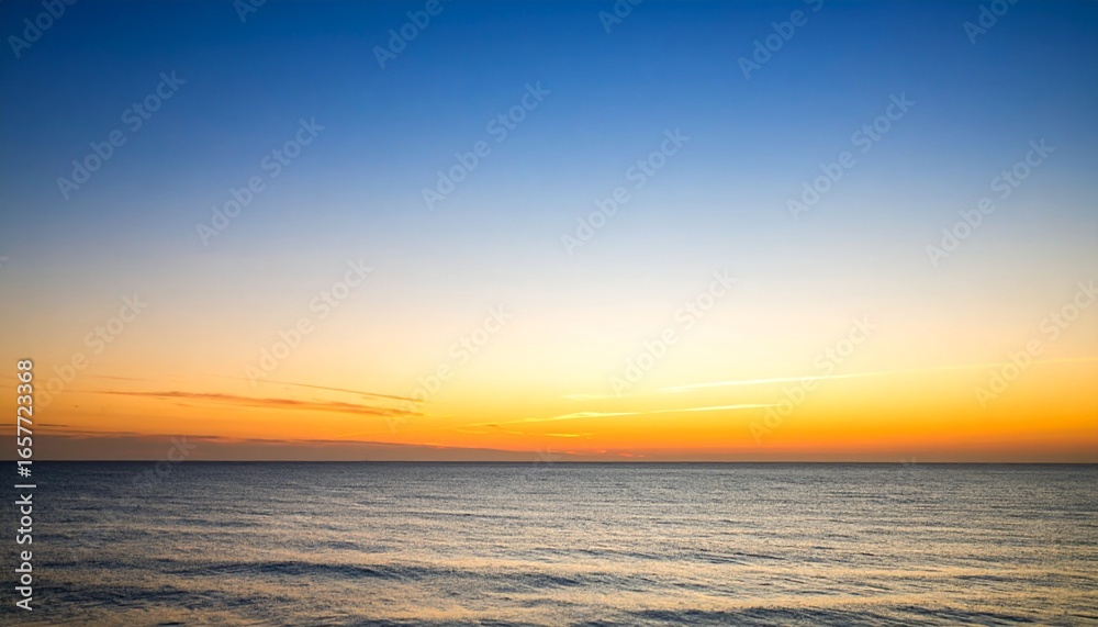 Obraz premium A serene seascape captures the transition from a vibrant orange horizon to a clear blue sky at sunset, reflecting light on the calm ocean surface.