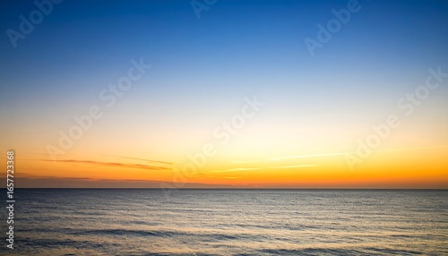 A serene seascape captures the transition from a vibrant orange horizon to a clear blue sky at sunset, reflecting light on the calm ocean surface.