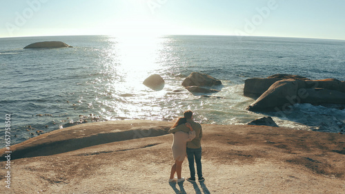 Hug, sunrise and back of couple by ocean for relaxing on holiday, vacation and weekend together. Dating, above and man with woman embrace by sea, water and rocks for bonding, relationship or romance