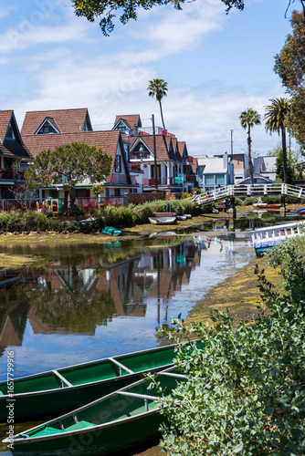 View of quaint houses with gabled roofs reflecting in the tranquil canal waters, as green boats rest along the banks in the vibrant sun, Los Angeles, California, United States.