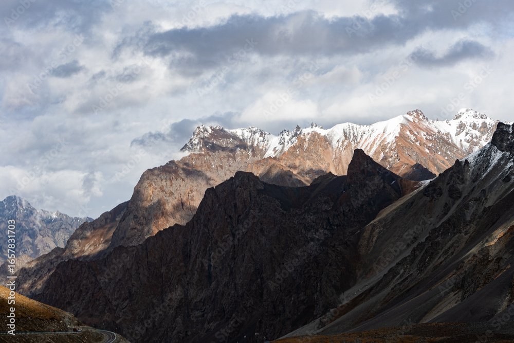 Obraz premium Dramatic sunlight breaks through clouds, casting a golden glow on a majestic mountain range while others remain in deep shadow. This high-contrast, epic landscape evokes power and awe.