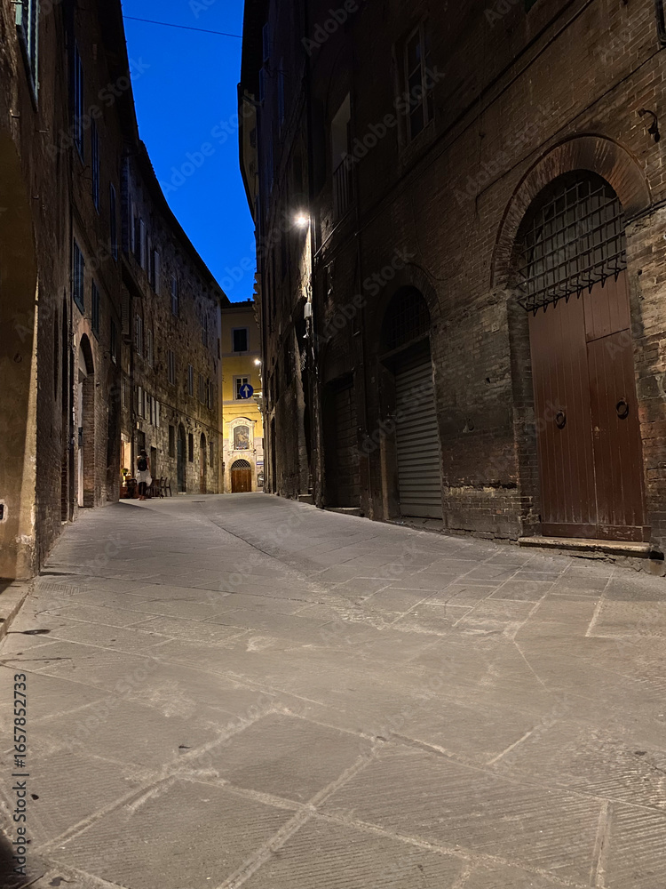 Fototapeta premium Narrow cobblestone street in Siena, Italy, illuminated by streetlights at dusk, creating a captivating blue hour atmosphere