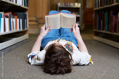 Female student reading book in a public library, lying on the ground