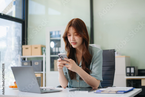 Asian woman, smiling and working at her desk in an office environment. She is wearing a business suit and holding documents