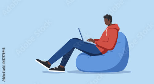 Young man in a red hoodie comfortably works on his laptop while sitting on a blue beanbag chair, depicting remote work, study, or online learning.