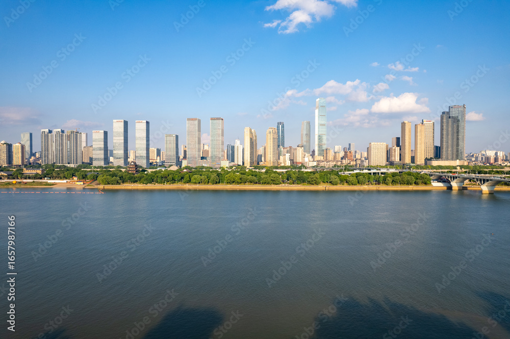 Naklejka premium Changsha City skyline in China displays modern buildings and a green riverfront reflecting in calm water under a bright blue sky.