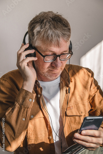 middle-aged man with glasses is enjoying music through his headphones while checking his smartphone. he is sitting in well-lit interior space, showing relaxed demeanor. close up.