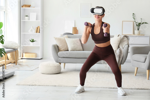 Sporty young woman in VR glasses exercising with dumbbells at home