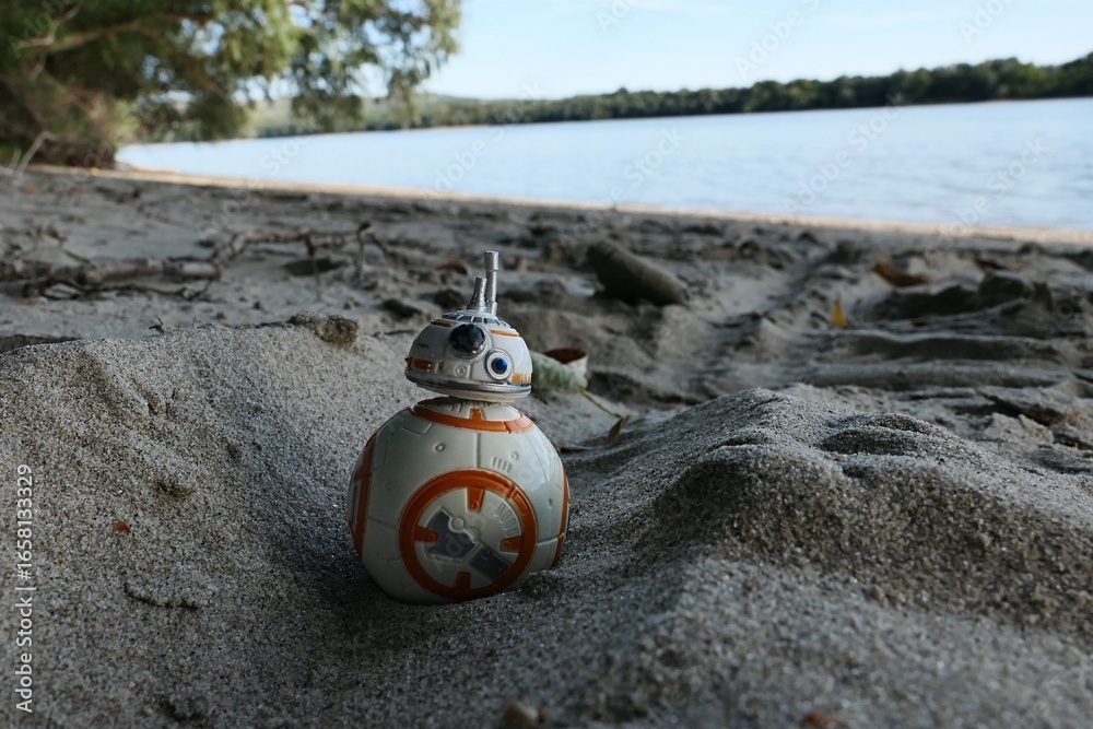 Fototapeta premium Star Wars figure of spherical rebel astromech droid BB-8 rolling in groove in sand leading from bank of Danube river. Riverside foliage, Danube river and blue skies in background. 