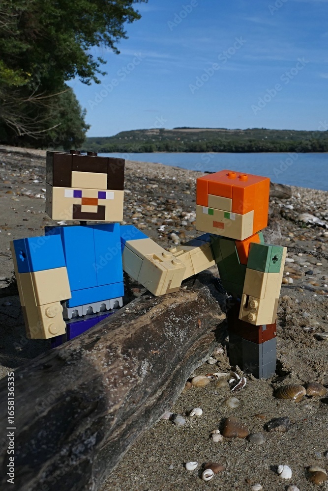 Fototapeta premium LEGO Minecraft action figures of Steve and Alex holding trunk of some driftwood on sandy bank of Danube river on borderline between southern Slovakia and Hungary. Summer blue skies, windblown clouds.