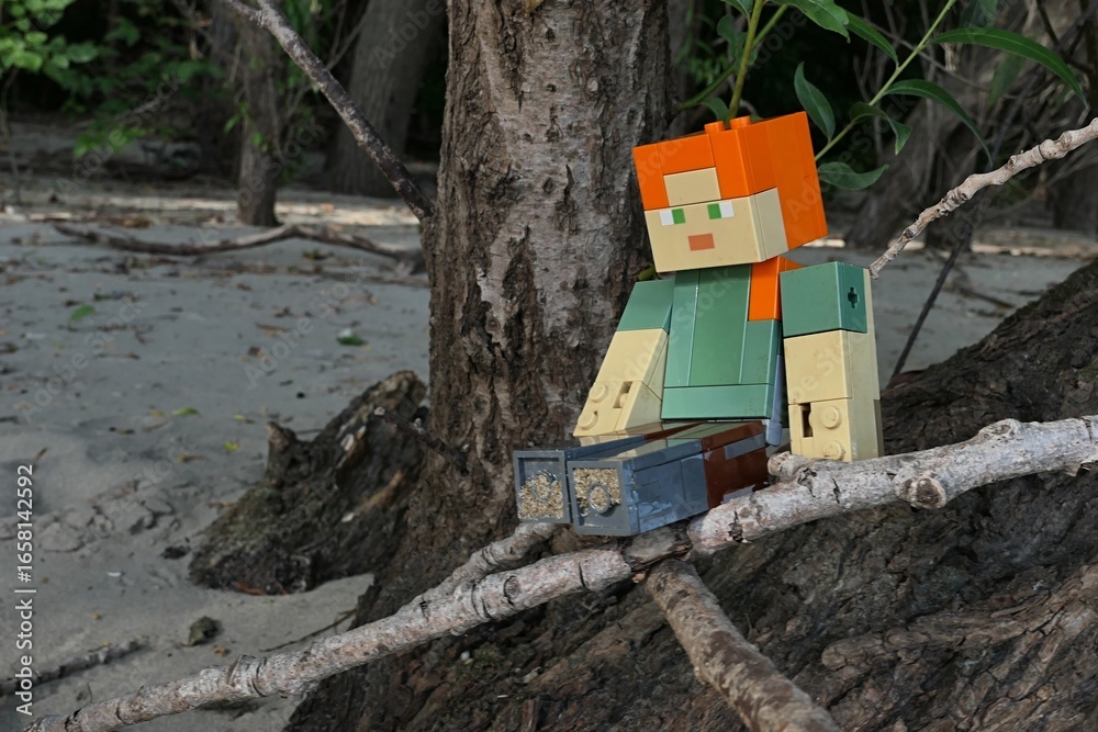 Obraz premium LEGO Minecraft action figure of main female character Alex, sitting on dried driftwood branch on sandy bank of Danube river, tree trunks and sandy beach in background.