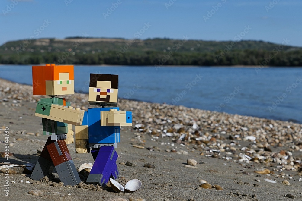 Obraz premium LEGO Minecraft figures of Steve and Alex, dancing waltz between clams and stones on sandy river Danube bank, borderline between Slovakia and Hungary. Forested opposite bank and blue skies visible.