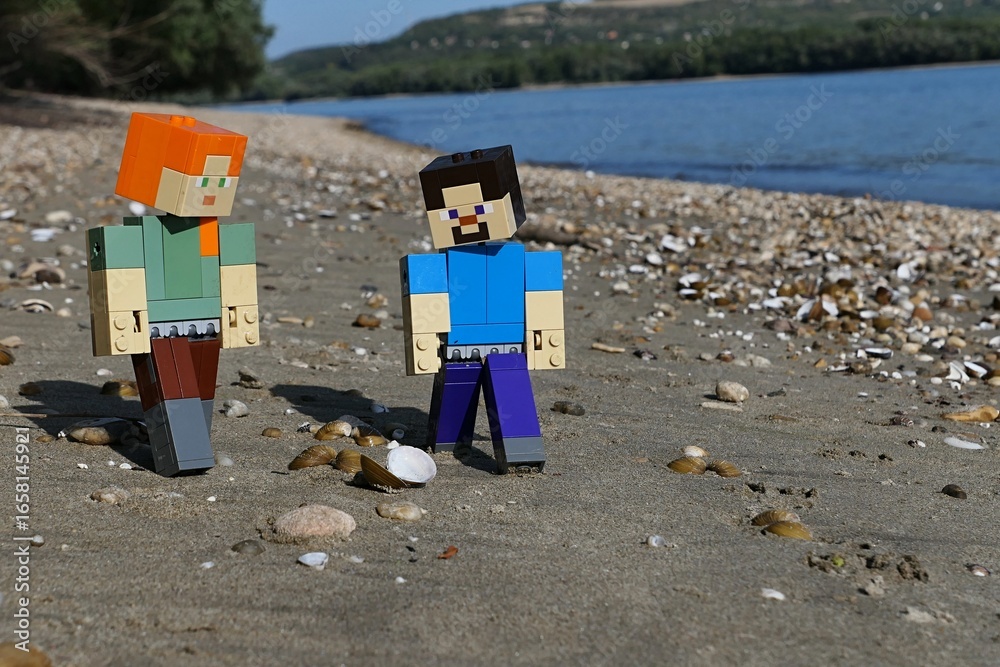 Obraz premium LEGO Minecraft action figures of Steve and Alex, examining clams and stones on sandy river Danube bank, borderline between Slovakia and Hungary. Forested opposite bank and blue skies visible.