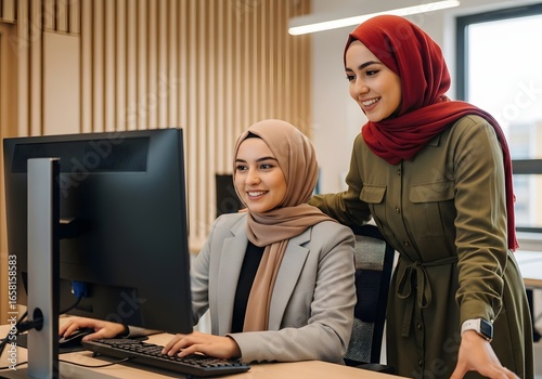 Two young muslim business women in hijab working together on a computer in a modern office, showcasing teamwork and collaboration