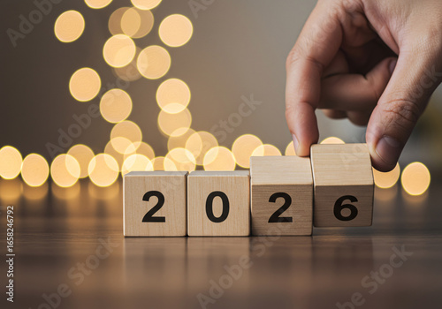 Close up of a hand placing the final wooden block to reveal the year 2026 with a bokeh background of golden lights