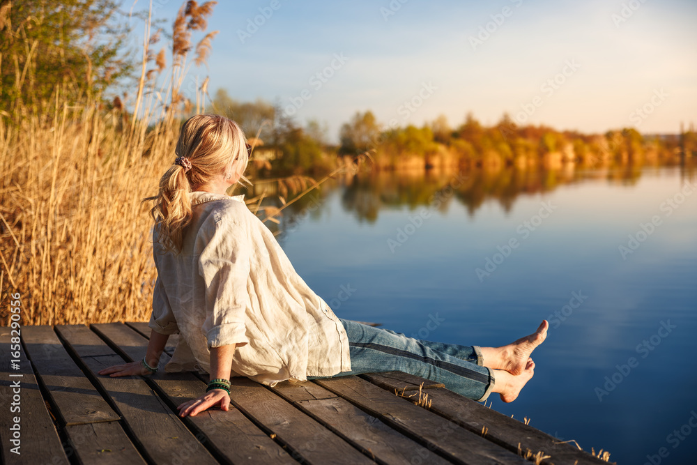 Obraz premium Woman sitting and relaxing barefoot on wooden pier by still lake during golden hour of indian summer