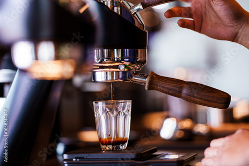 Close-up of a barista preparing espresso with a professional coffee machine. Fresh coffee is being extracted into a clear glass placed on a digital scale. The warm lighting and rich crema highlight th