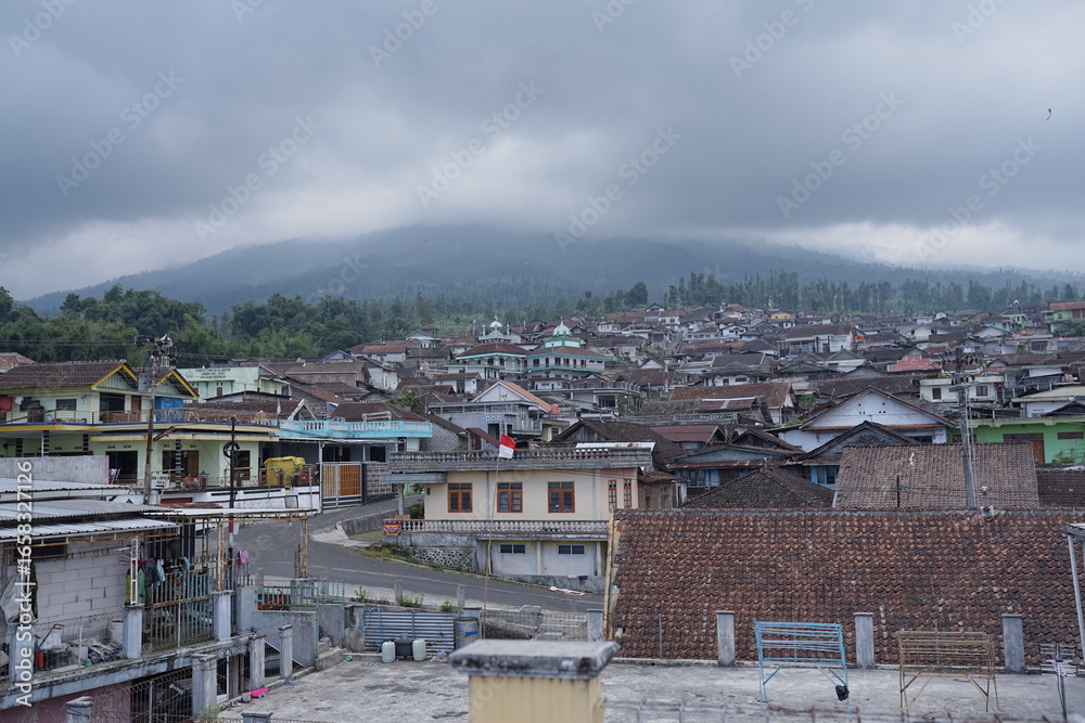 Fototapeta premium Sunday, August 3, 2025, 14:19:44, Temanggung, Indonesia _ Panoramic view of a densely populated village nestled at the foot of a majestic, cloud-covered mountain.