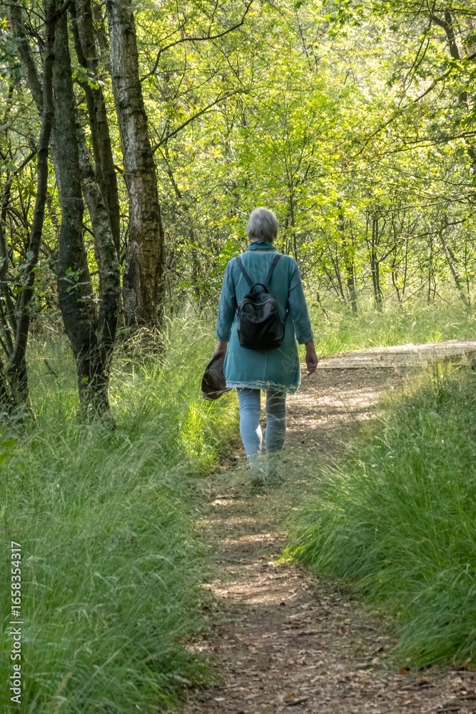 Obraz premium Senior woman walking on path in green forest holding hat and backpack