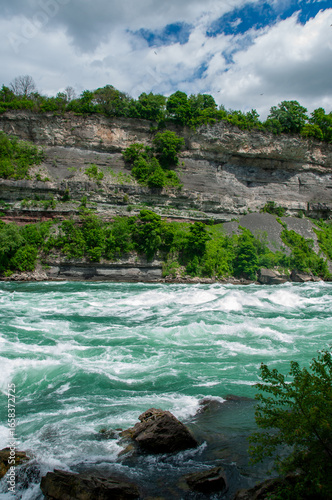 The powerful, turquoise rapids of the Niagara River flowing through a lush green gorge with rocky cliffs. The sky is partly cloudy, and the trees are vibrant green, suggesting a warm and sunny day.