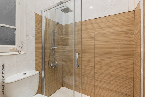 Modern bathroom with a glass shower enclosure and wooden wall panels. White toilet and tiled floor are visible. Bright lighting enhances the clean design.
