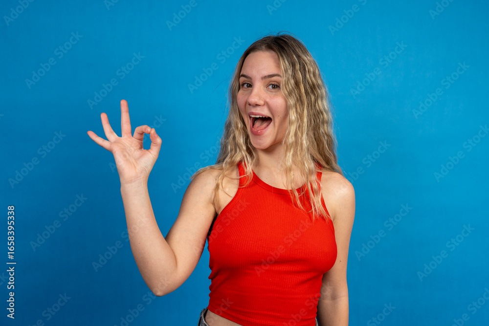 Fototapeta premium Enthusiastic young woman showing ok sign with blue background